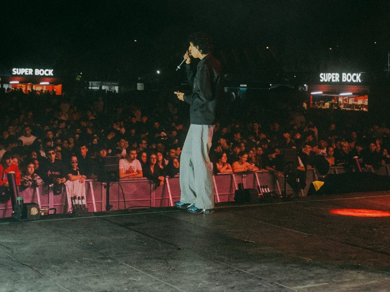 A performer on stage facing a packed crowd at night, festival bars lit in the background