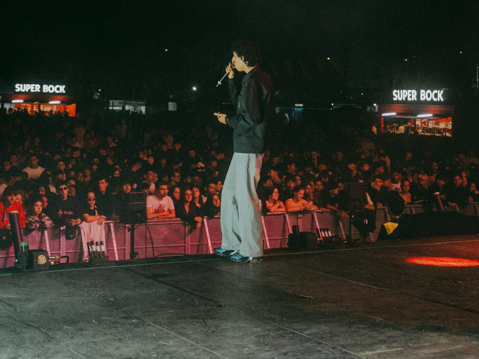 A performer on stage facing a packed crowd at night, festival bars lit in the background